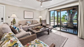 A spacious living room with a sofa, armchairs, and a coffee table. There is a ceiling fan, a framed artwork on the wall, and a view of the outdoors through large glass doors.