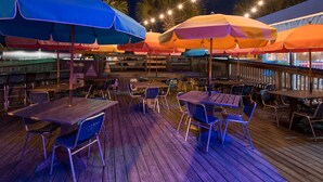 Un espace extérieur aménagé pour les repas, avec des tables et des chaises en bois, protégé par des parasols colorés, et illuminé par des guirlandes lumineuses en arrière-plan.