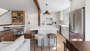 Una cocina moderna con armarios blancos, una isla central, taburetes de bar, una zona de comedor con mesa y sillas, y una lámpara colgante.