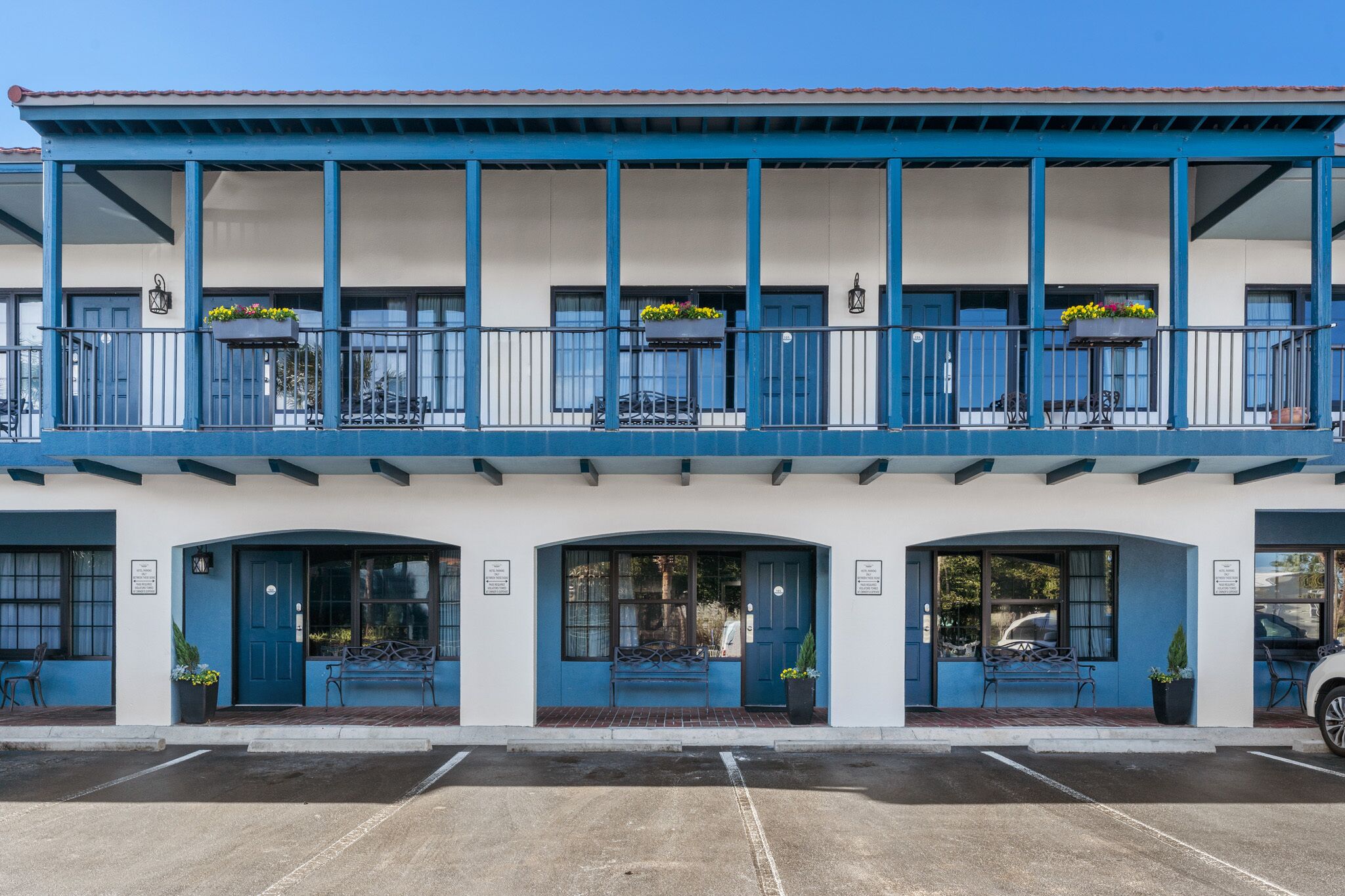 Un immeuble à la façade bleue et blanche, doté de nombreux balcons ornés de jardinières fleuries, et un parking situé devant.