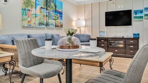 A dining area with a round table, grey upholstered chairs, a glass-top table, a ceiling fan, and a wall-mounted flat-screen TV. - 2 Queen Beds. Don`t Leave Your Dog at Home! (Destin)