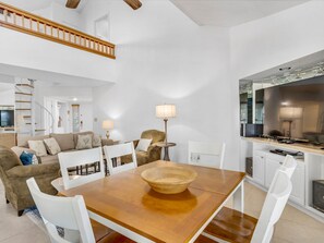 A spacious living area with a wooden dining table, white chairs, a sofa, and a TV.