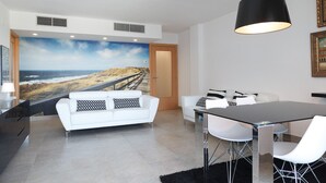 Een moderne woonkamer met een grote muurschildering van uitzicht op de oceaan, witte banken, een eettafel met stoelen en een zwarte hanglamp.
