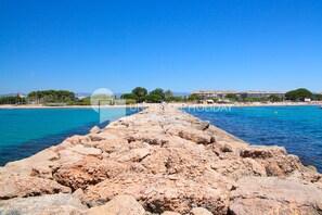 Un muelle rocoso que conduce a una playa con agua azul cristalina, palmeras y edificios al fondo.