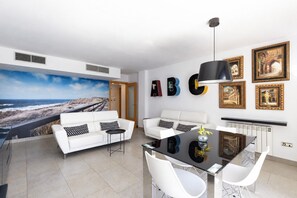 En moderne stue med et stort mural af et kystlandskab, to hvide sofaer med mønstrede puder, et glas spisebord med stole og en sort pendel.