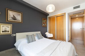 A hotel room with a bed, a nightstand, a wall-mounted lamp, and a door. - Spacious family apartment with sea views and pool · UHC MAR AUGUSTA 083 (Cambrils)