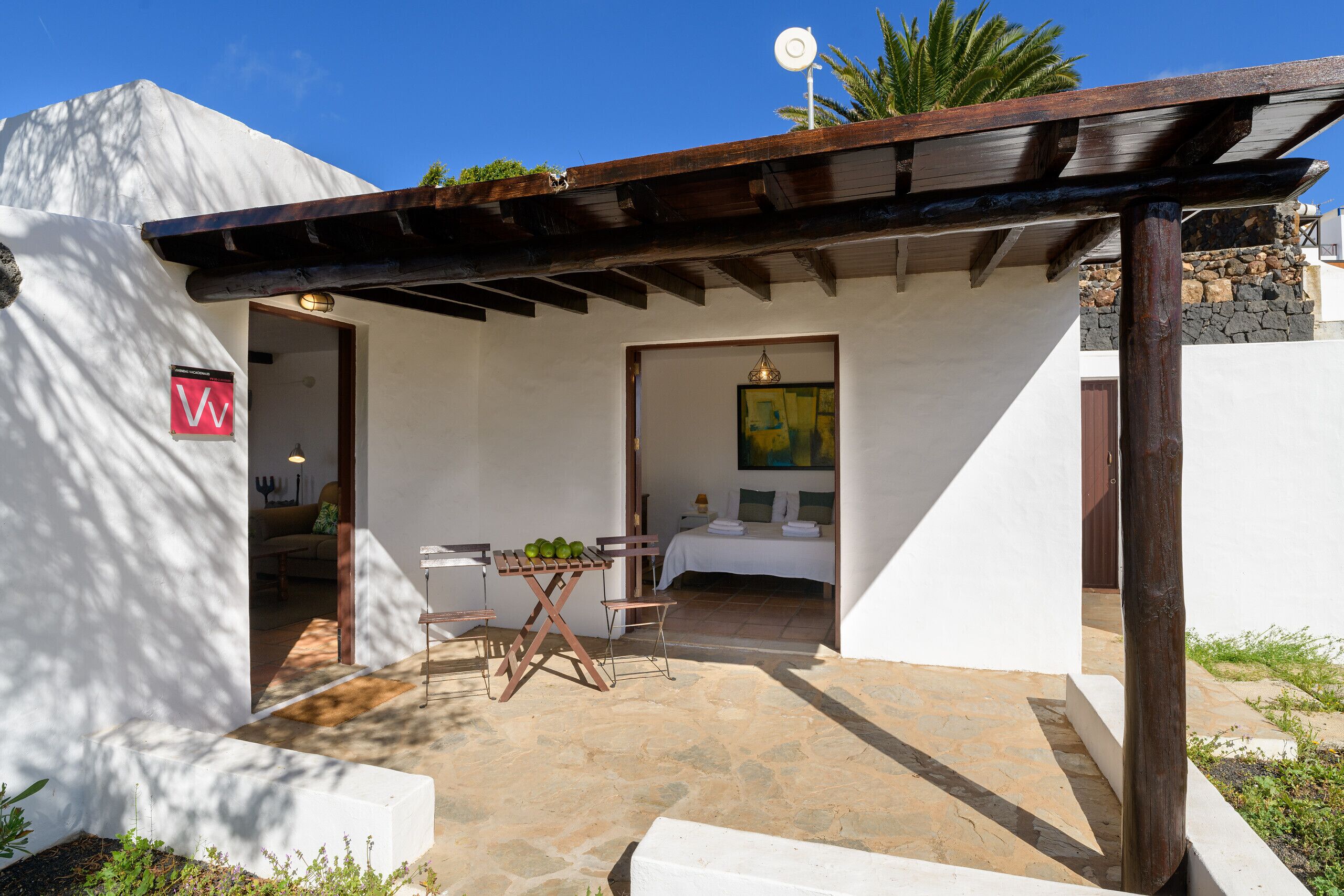Un pequeño edificio blanco con un patio de piedra, una pérgola de madera y una cama con un cuadro.