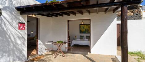 Un pequeño edificio blanco con un patio de piedra, una pérgola de madera y una cama con un cuadro.
