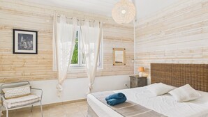 Ein Schlafzimmer mit einem Bett, einem Sessel, einem Nachttisch, einer Lampe und einem Fenster mit Vorhängen.