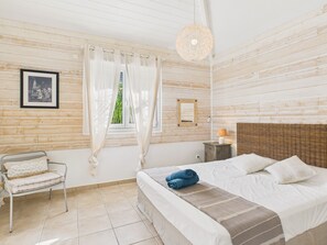 Ein Schlafzimmer mit einem Bett, einem Sessel, einem Nachttisch, einer Lampe und einem Fenster mit Vorhängen.