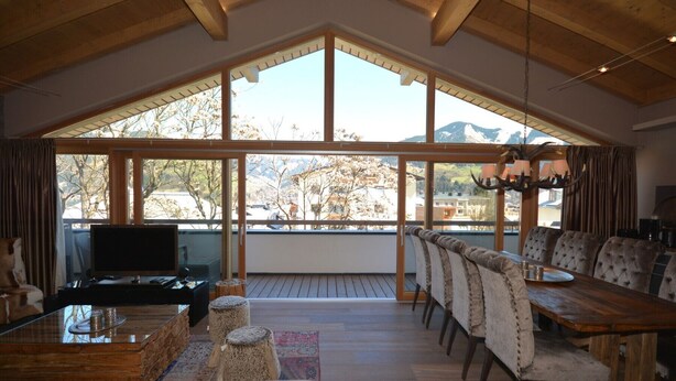 A spacious living area with a high ceiling, a large window offering a mountain view, a dining table with chairs, and a television on a wooden coffee table.