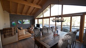 A spacious living area with a wooden ceiling, large windows, a dining table with upholstered chairs, and a fireplace.