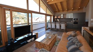A modern living room with a large TV, wooden furniture, and a balcony with a view of the mountains.