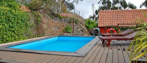 Una piscina con cubierta de madera, rodeada de vegetación y un edificio de techo rojo.