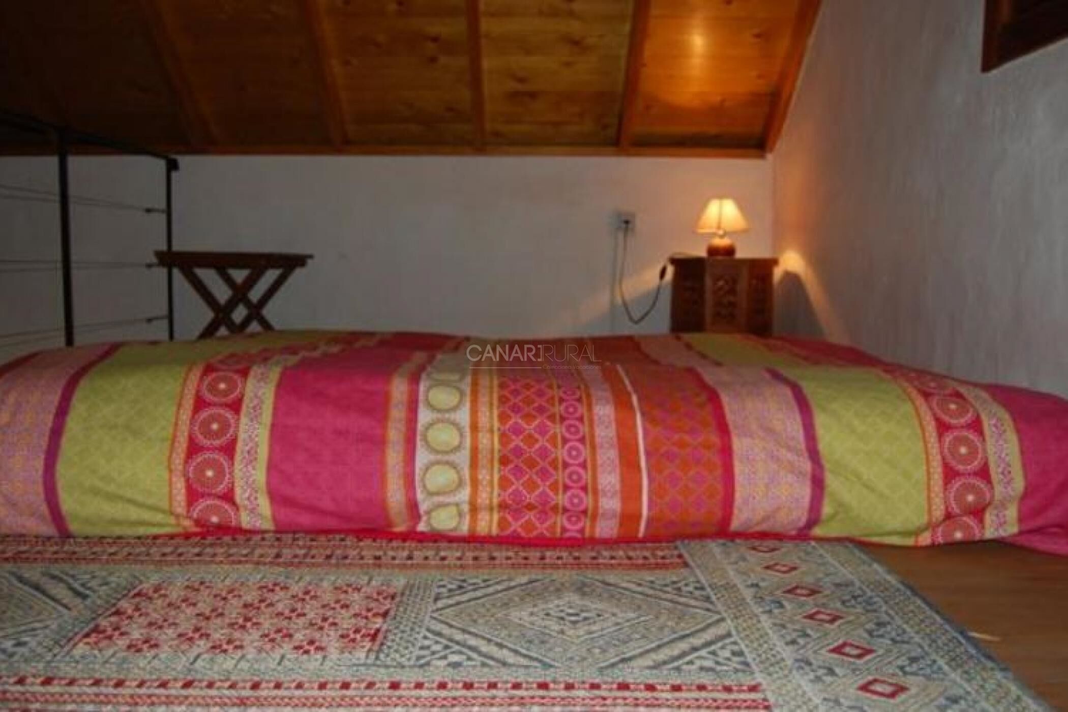 Un dormitorio con una cama, techo de madera, una lámpara de pared y una mesita.