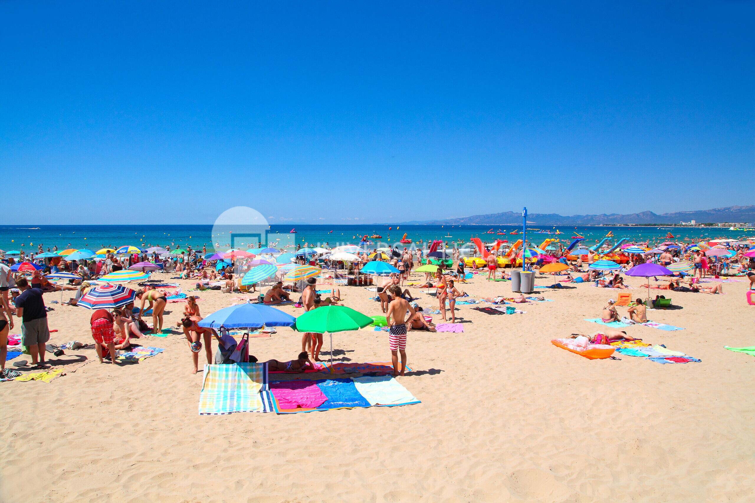 Ein belebter Strand mit badenden und sonnendenden Menschen, bunten Sonnenschirmen und einem strahlend blauen Himmel.