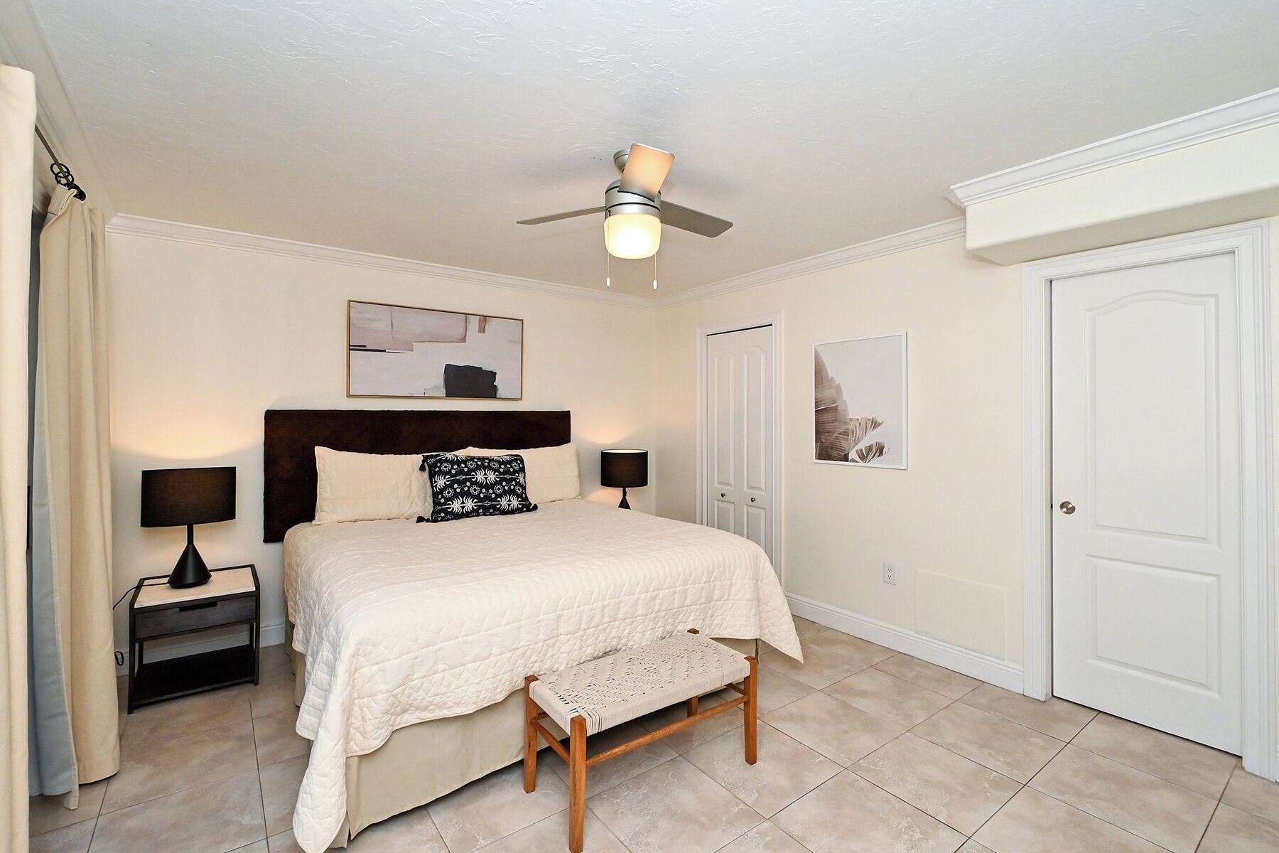 A bedroom with a bed, bedside tables, a bench, a ceiling fan, and two doors.