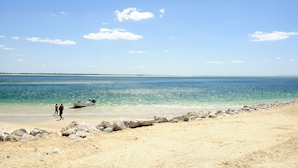 A beach with clear blue water, a rocky shoreline, and a few people walking along the shore. - Refurbished apartment with air conditioning in the center of Setúbal (Setúbal)