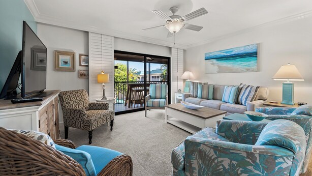 Un salon avec un ventilateur de plafond, un téléviseur, un canapé, un fauteuil et un tableau représentant une scène de plage.