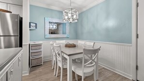 Un coin repas avec une table en bois, des chaises blanches, un luminaire suspendu et un réfrigérateur.