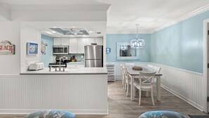 Une cuisine moderne avec des meubles blancs, un îlot central et un coin repas agrémenté d’un mur bleu clair et de chaises blanches.