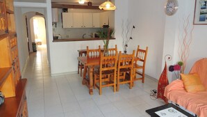 A dining area with a wooden table and chairs, a kitchen in the background, and a living area with a sofa and a clock on the wall.