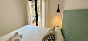 Un lit bien fait, avec du linge de lit blanc et des serviettes pliées, une tête de lit verte et une petite plante en pot sur la table de chevet.