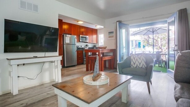 A modern living room with a flat-screen TV, a wooden coffee table, and a blue armchair. A kitchen with wooden cabinets and a dining area with a table and chairs is visible through an open door. - Newly Renovated Condo - Close to Everything! (San Diego)