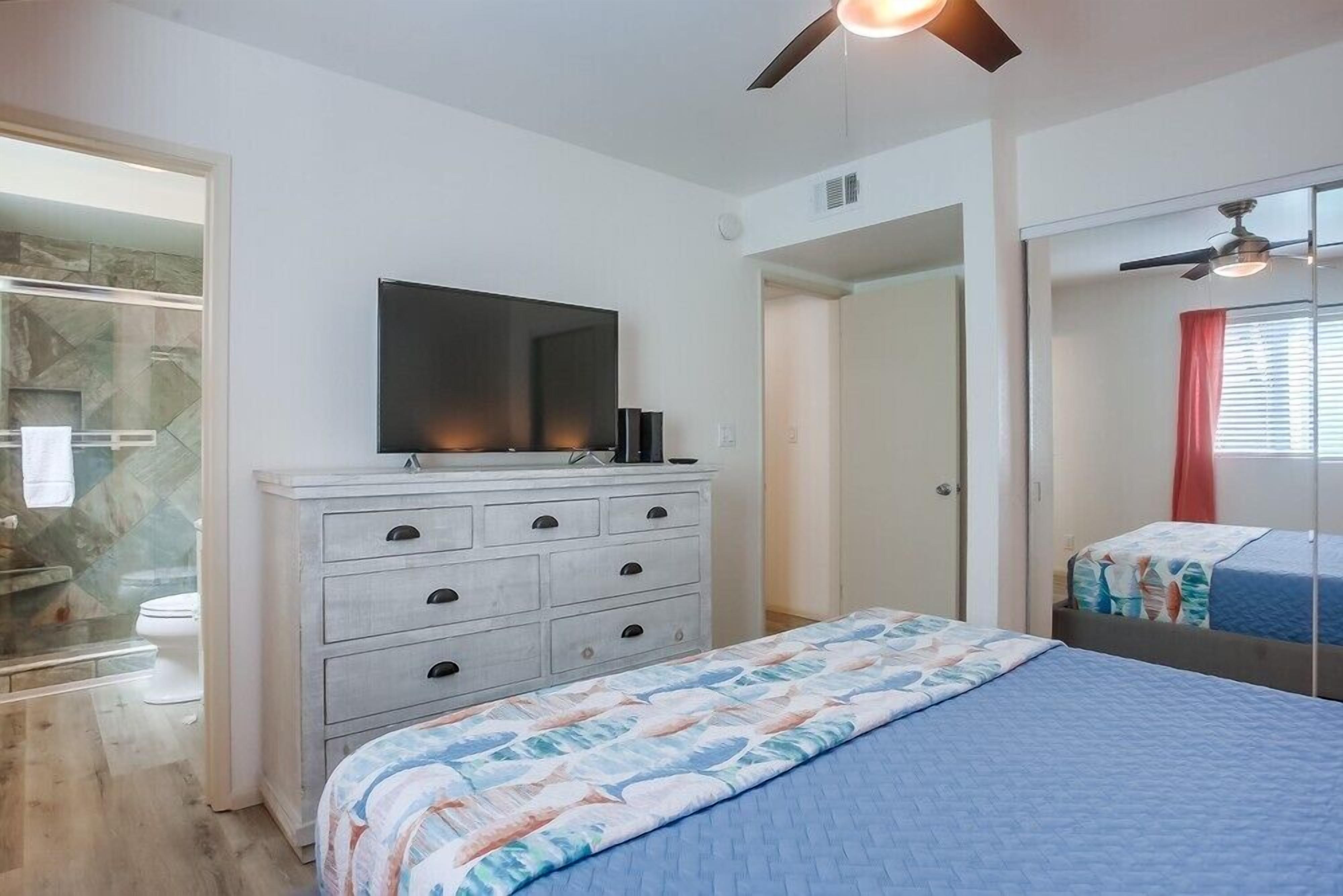A bedroom with a bed, a dresser, a television, and a bathroom visible through an open door.
