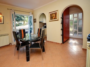 Un coin repas avec une table et des chaises en verre, une cuisine à l’arrière-plan et une porte rouge donnant sur l’extérieur.