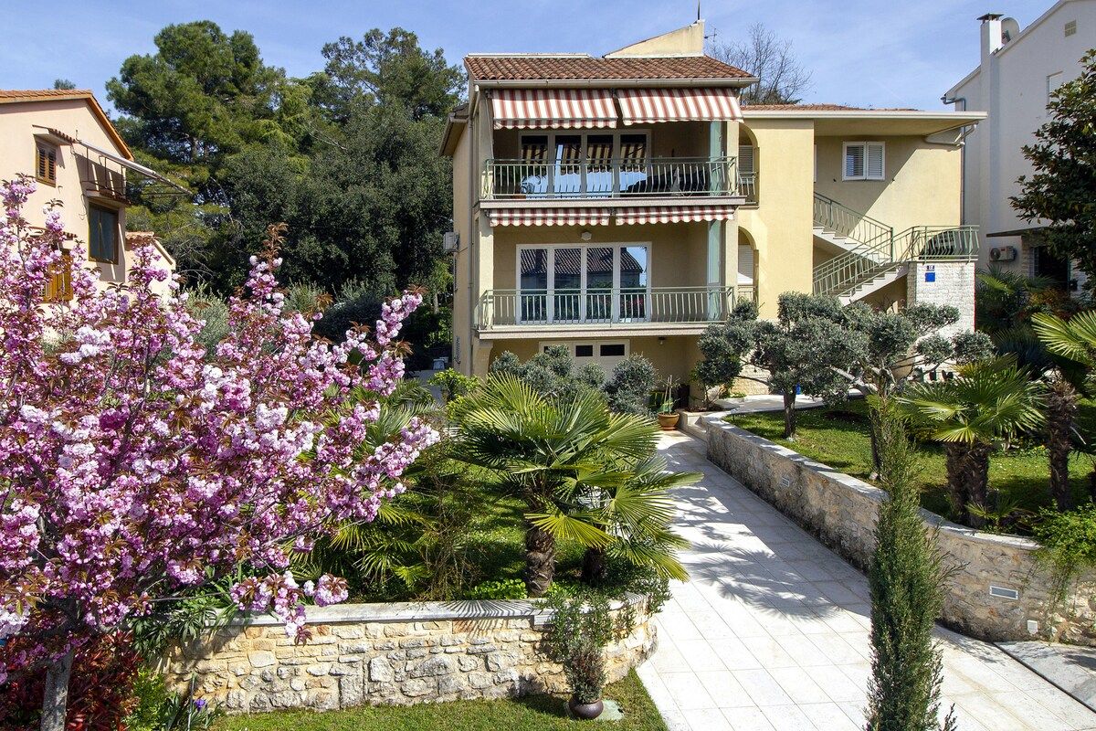 A residential building with a balcony, a stone pathway, and surrounding greenery.