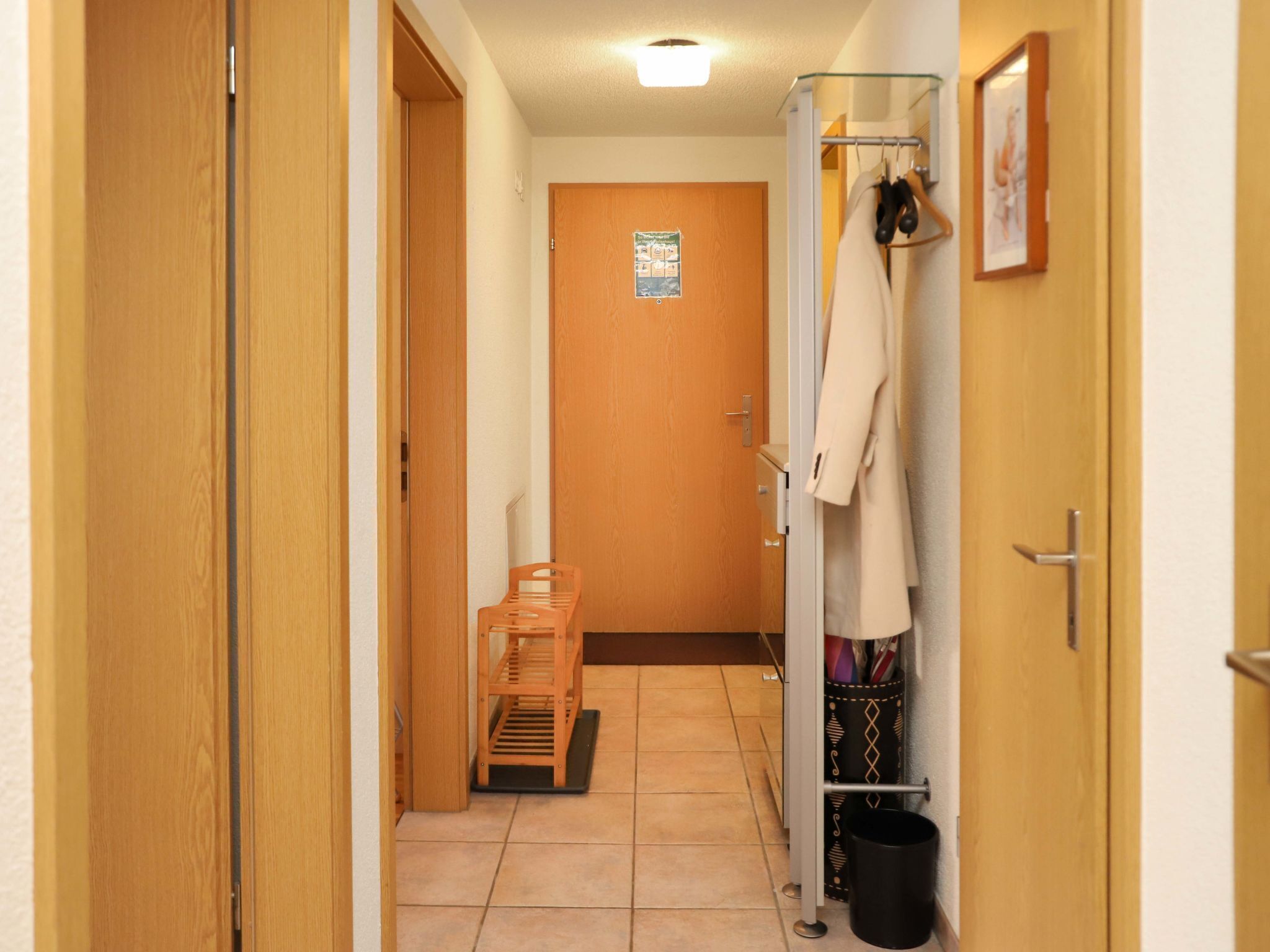 Un couloir avec des portes en bois, un porte-manteau sur lequel est accroché un manteau, un banc et une poubelle.