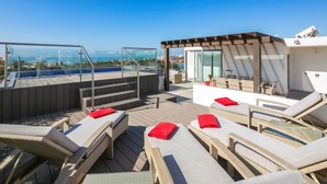 A rooftop terrace with outdoor seating, a glass balustrade, and a view of the sea.