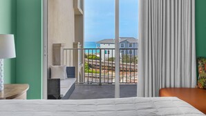 Quarto de hotel com cama, mesa de cabeceira, luminária e varanda com vista para o oceano e outros edifícios.