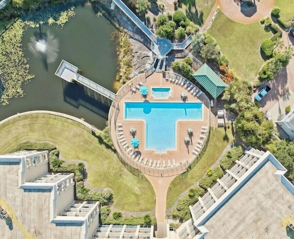 An aerial view of a hotel with a central swimming pool, a bridge, and surrounding greenery.