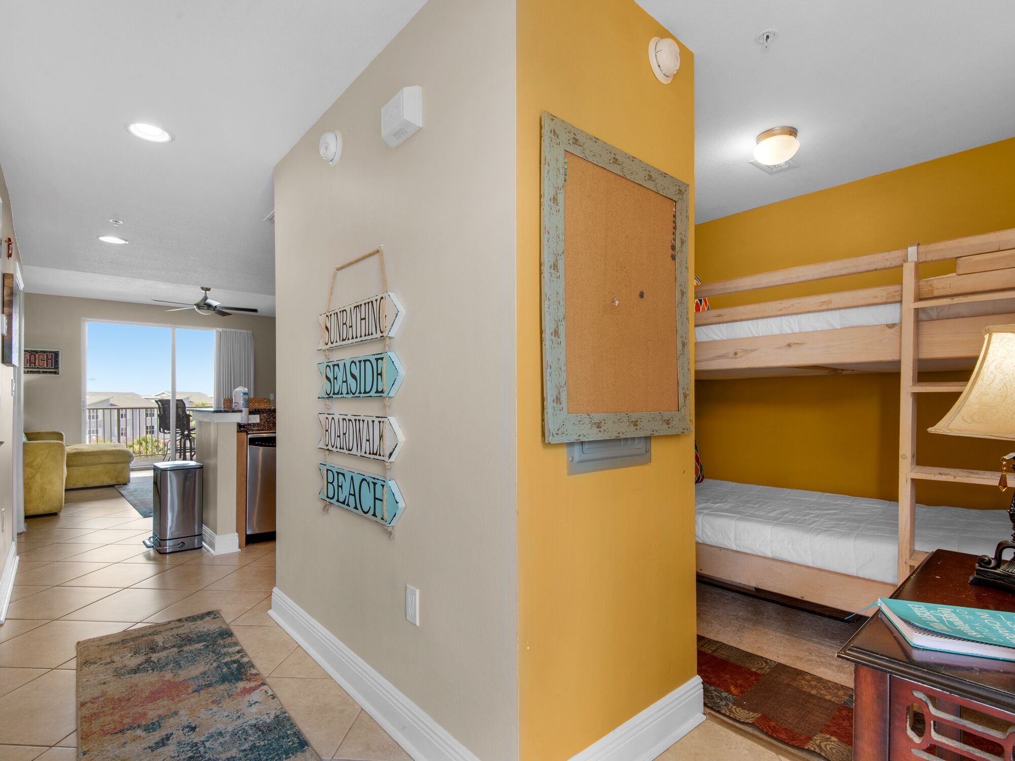 A room with a bunk bed, a corkboard with directional signs, a kitchen area, and a view to the outside.
