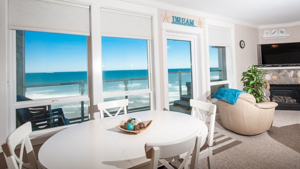 Een woonkamer met een witte tafel en stoelen, een beige bank, een televisie en een open haard. Grote ramen bieden uitzicht op de oceaan.