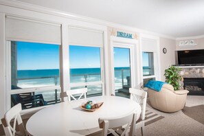 Un salón con una mesa y sillas blancas, un sofá beige, un televisor y una chimenea. Amplias ventanas ofrecen vistas al océano.
