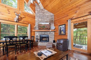 A wooden cabin interior with a stone fireplace, dining area, and a view of the outdoors.