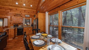 Eine Holzhütte mit Essbereich, Küche und großen Fenstern mit Blick auf einen Wald.