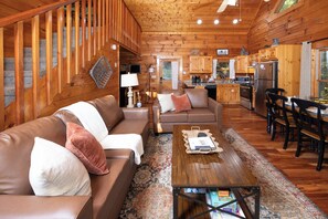 A cozy cabin interior with a wooden staircase, a living room with a sofa and armchairs, a dining area with a table and chairs, and a kitchen with wooden cabinets and appliances.