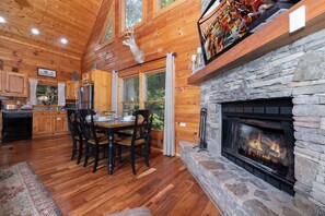 A wooden cabin interior with a stone fireplace, dining table, chairs, and a kitchen area. - Pool Table, Hot Tub, Arcade Games!! Pool, Putt Putt, Fishing, Golf! (Sevierville)