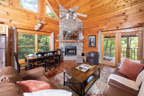 A cabin interior with a stone fireplace, wooden walls, a dining area, and a large window overlooking greenery.