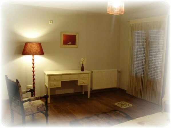 A room with a wooden floor, a white radiator, a desk with drawers, a lamp, a chair, and a window with blinds.