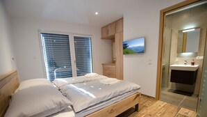 A bedroom with a wooden bed, a large window with blinds, a wooden wardrobe, a mounted TV, and a bathroom with a glass shower enclosure.