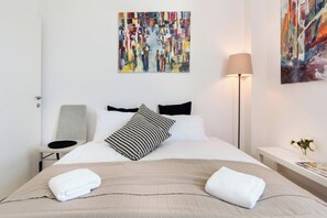 A hotel room with a bed, a chair, a lamp, and two paintings on the wall.