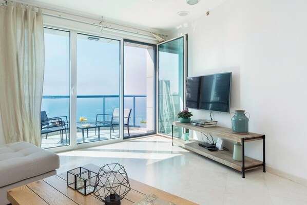 Una sala de estar moderna con un ventanal corredero que da a un balcón, un televisor de pantalla plana montado en la pared y una mesa de centro de madera con objetos decorativos.