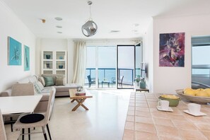 Una sala moderna con sofá, mesa de comedor y sillas, un balcón con vistas, y un área de cocina con frutero y tazas.
