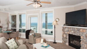 A living room with a fireplace, large windows overlooking the ocean, a ceiling fan, and a flat-screen TV. - Promo! Oceanfront Condo - Private Hot Tub, WiFi, HDTV and Pool! (Lincoln City)