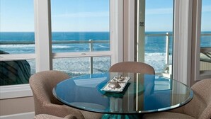 A round glass table with a plate of food and two chairs, overlooking a beach with waves and a clear sky. - Promo! Oceanfront Condo - Private Hot Tub, WiFi, HDTV and Pool! (Lincoln City)